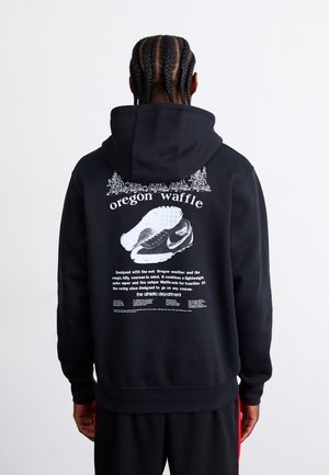 Osoba z warkoczami nosi czarną bluzę z kapturem z napisem „Oregon Waffle” i grafiką butów sportowych na plecach, zwrócona tyłem do kamery.