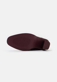 Suola di scarpa bordeaux con un grip testurizzato, caratterizzata da un tacco quadrato e da leggere scanalature per la trazione. Finitura liscia e opaca senza loghi visibili.