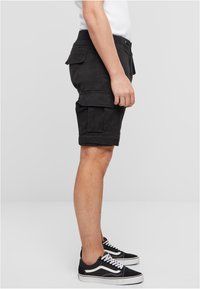 Juodi krovinių šortai su šoninėmis kišenėmis, tiesiu kirpimu ir apačia šiek tiek aukščiau kelio, derinami su juodai baltais sportbačiais.