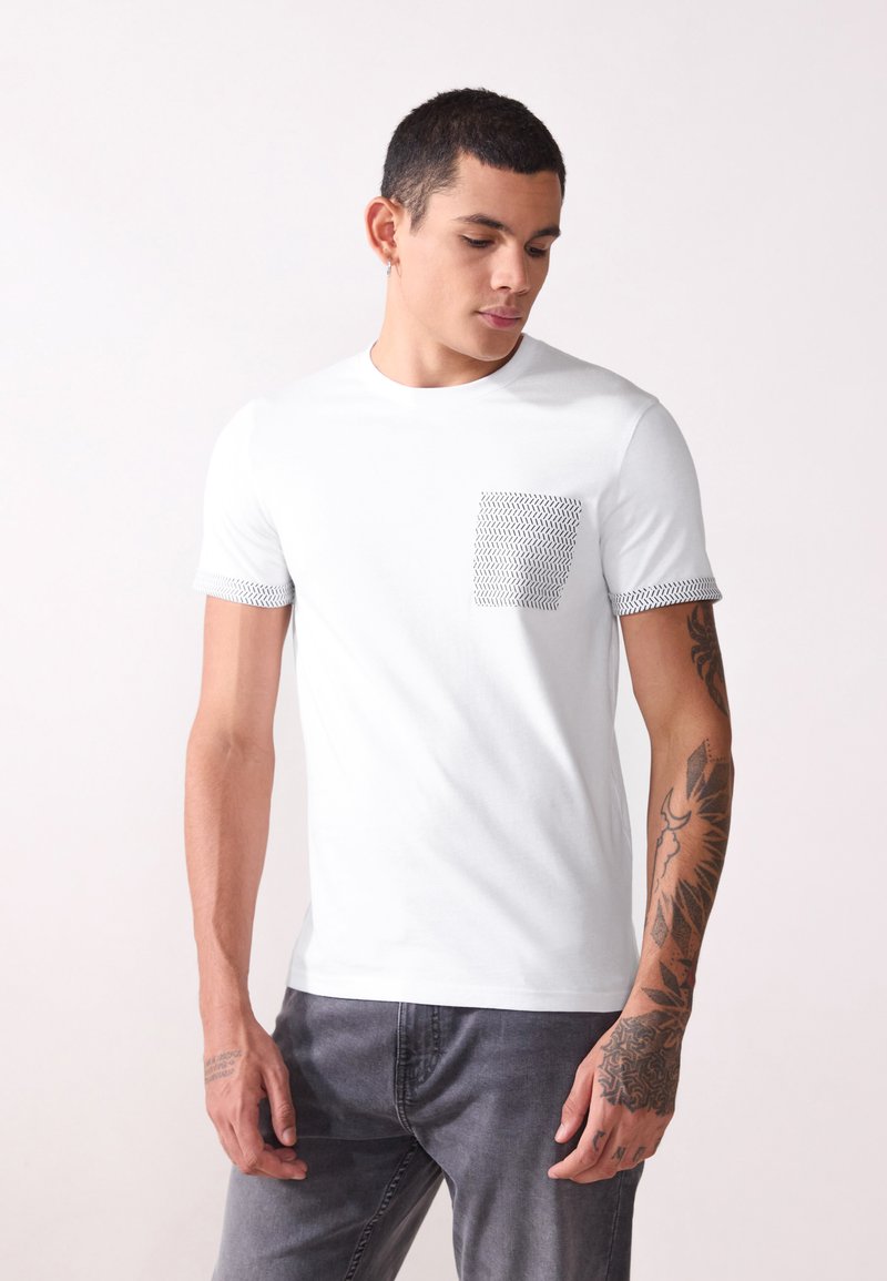 Valkoinen lyhythihainen t-paita, joka on valmistettu puuvillasta. T-paidassa on kuviollinen tasku ja teksturoidut hihat. Slim fit -muotoilu, jossa on pyöreä kaula-aukko.