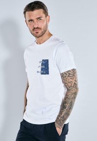 White cotton t-shirt with short sleeves, featuring a blue textured rectangular patch with "EDGE OF THE HORIZON" text and a floral pattern.
