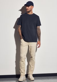 Camiseta de cuello redondo azul marino, pantalones tapered beige y zapatillas ligeras. Presenta tatuajes en los brazos y una gorra de béisbol azul marino a juego.