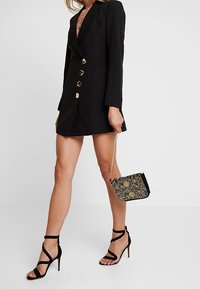 Sort skræddersyet blazer-kjole med guldknapper, kombineret med sorte højhælede sandaler med stropper og en lille sort og guldfarvet mønstret taske.