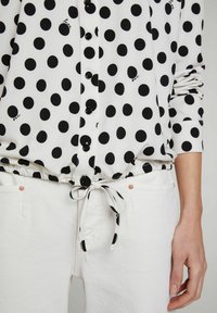Camicia nera a pois con chiusura a bottoni, orlo elasticizzato e pantaloni bianchi con vita regolabile e dettagli metallici. Tessuto morbido.