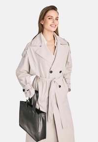 Beżowy trencz o podwójnym zapięciu, z dużymi klapami i przewiązanym paskiem w talii. Czarna skórzana torba na ramię z krótkimi uchwytami.