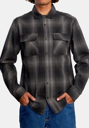 Mężczyzna ubrany w czarno-szarą koszulę w kratę z kieszeniami na piersi i niebieskie dżinsy, ręce w kieszeniach.