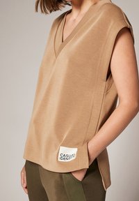 Camelowy top z krótkim rękawem i dekoltem w kształcie litery V, z boczną kieszenią i metką. Wykonany z miękkiego, teksturowanego materiału.
