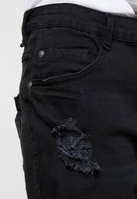 Czarne dżinsy z efektem przetarcia na udzie, z postrzępionymi brzegami i metalowymi guzikami w kieszeniach.