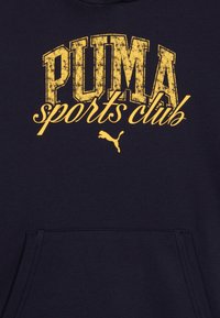 Sweatshirt bleu marine avec le texte jaune usé "PUMA sports club" et un petit logo Puma noir au-dessus de la poche avant. Tissu texturé.