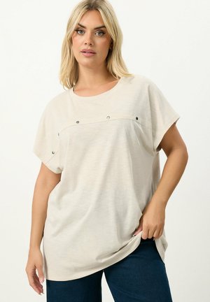 Kvinde iført løs beige kortærmet top med metalnitter hen over brystet og mørkeblå bukser, stående mod en ensfarvet hvid baggrund.