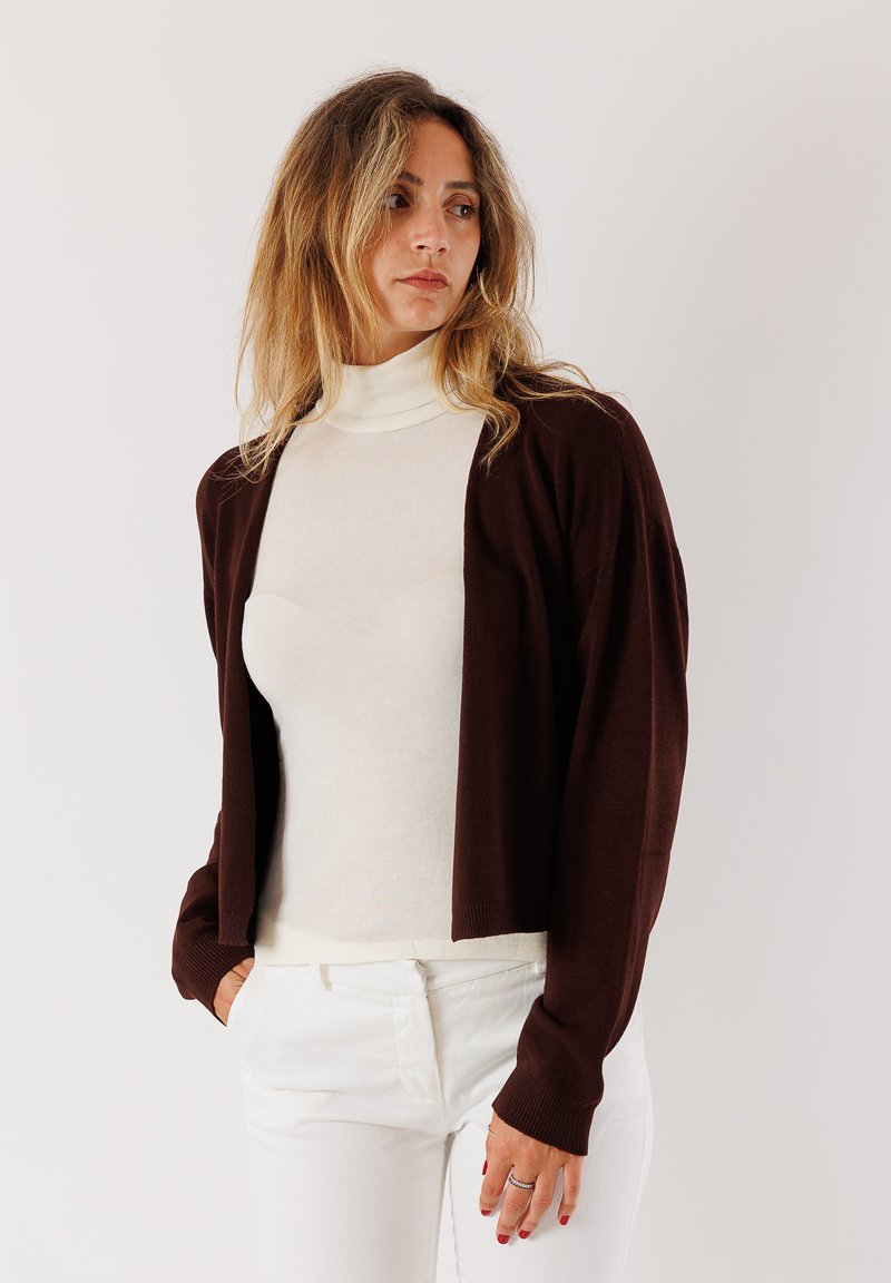 Donna con capelli biondi ondulati che indossa un cardigan marrone scuro sopra un dolcevita bianco e pantaloni bianchi, in piedi contro uno sfondo chiaro e uniforme.