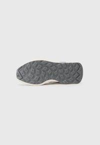 Semelle de chaussure avec un fond en caoutchouc noir texturé et un côté crème, présentant un motif géométrique et un détail de logo en relief.