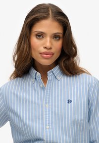 Jasnoniebieska koszula na guziki z białymi pionowymi paskami. Materiał bawełniany z małym haftowanym logo na lewej piersi. Standardowy kołnierz.