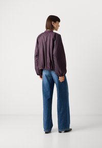 Giacca bomber viola con orlo elasticizzato, abbinata a jeans in denim blu a gamba larga. Texture liscia; i dettagli includono polsi e colletto a coste.