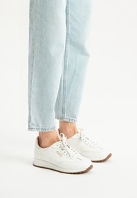 Sneakers bianchi in pelle con finitura testurizzata, design con lacci e suola in gomma marrone; presenta un logo dorato sul lato.