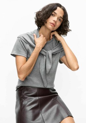 Vrouw met krullend haar, een grijs T-shirt met korte mouwen en een leren rok dragend, die een grijze trui die over haar schouders is gedrapeerd, rechtzet.