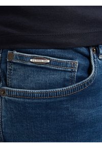 Vaqueros de mezclilla en azul medio con una textura desgastada, con una etiqueta plateada "PETROLIND" en el bolsillo delantero y costuras en contraste.