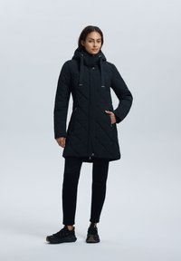Zwarte gewatteerde lange jas met een capuchon, voorzien van een rits aan de voorkant, trekkoorden en zijzakken. Gedragen met zwarte broek en sneakers.