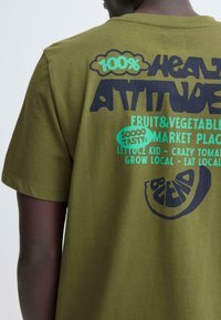 Olijfgroen T-shirt van katoen met gedurfde grafische ontwerpen in donkerblauw en aqua, met fruit- en groentethema's op de achterkant.