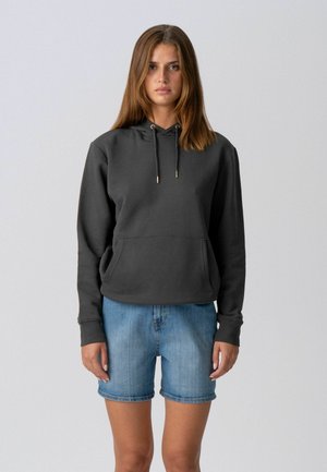 Junge Frau mit glatten braunen Haaren, die einen schwarzen Hoodie und helle Jeansshorts trägt, vor einem schlichten hellen Hintergrund stehend.