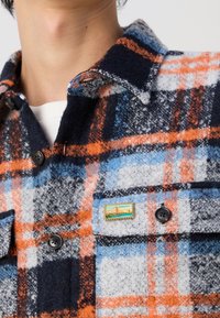 Flanelová košile s modro-oranžovo-šedým kostkovaným vzorem. Disponuje límečkem na knoflíky, černými knoflíky a dekoračním špendlíkem na náprsní kapse.