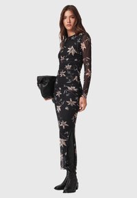 Lång svart klänning med blommönster, genomskinliga långa ärmar, sprund på sidan och figurnära design. Tillsammans med en svart handväska och ankelboots.