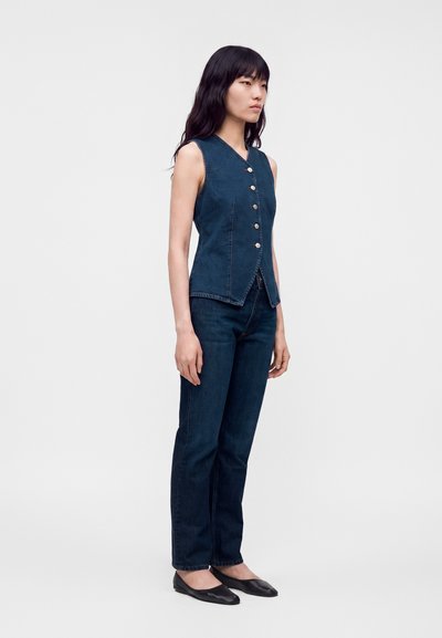 Femme aux longs cheveux noirs portant un gilet en denim bleu foncé sans manches boutonné, un jean assorti et des ballerines noires, debout devant un fond uni.