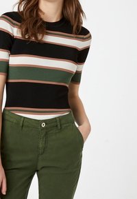Musta ja raidallinen lyhythihainen paita, jossa on vihreitä, valkoisia ja ruosteisia kuvioita, yhdistettynä oliivinvihreisiin housuihin, joissa on etutasku ja napitus.