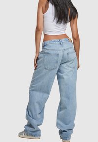 Jean en denim bleu clair, ample, taille haute, avec deux poches arrière et une étiquette en cuir beige à la ceinture.