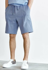 Shorts de sport bleu clair avec une taille à cordon, des poches avant et une texture lisse, associés à des baskets blanches avec des accents gris.