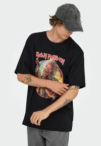 Camiseta de algodón negra con un gráfico vibrante de un monstruo con pelo espigado y un hacha, con la marca "Iron Maiden" en letras rojas negritas.