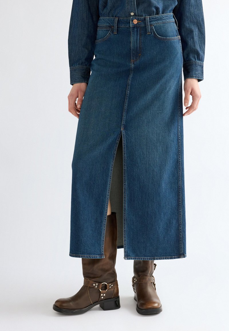 Femme portant une longue jupe en jean bleue avec une fente frontale, associée à des bottines en cuir marron sur un fond clair uni.