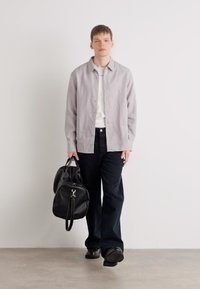 Grijze overhemd met knopen over een wit T-shirt, gecombineerd met donkerblauwe broek. Model houdt een zwarte leren tas vast en draagt zwarte schoenen.