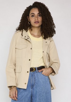 Donna con capelli ricci indossa una giacca beige, un maglione giallo chiaro, jeans blu e orecchini a cerchio dorati, posando con una mano in tasca.