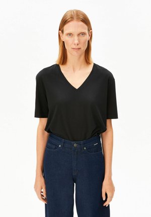 Vrouw met recht rood haar draagt een zwart T-shirt met korte mouwen en V-hals, ingestopt in een high-waist donkerblauwe spijkerbroek, staand tegen een effen witte achtergrond.