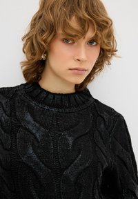 Schwarzer Strickpullover mit hohem Ausschnitt, strukturiertem Design und gerippten Akzenten. Das Modell hat gestuftes, welliges Haar und minimalen Schmuck.
