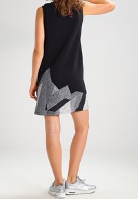 Ärmlös svart klänning med geometriska grå mönster vid fållen, gjord av mjukt tyg. Bärs med silverfärgade träningsskor.