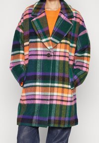 Person iført en flerfarvet ternet frakke med grønne, orange, pink og lilla nuancer over en orange sweater, hænderne i lommerne.