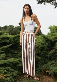 Jeune femme aux longs cheveux foncés portant un débardeur blanc, un pantalon rayé vertical marron et blanc, et des sandales, debout à l'extérieur parmi des buissons verts.