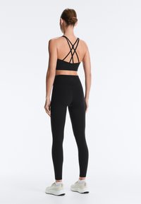 Žena s vlasy staženými do drdolu, oblečená v černém sportovním topu s ramínky, černých legínách s vysokým pasem a bílých teniskách, otočená zády na jednoduchém pozadí.