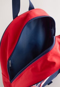 Red and navy backpack with a smooth, durable exterior. Interior is lined in blue fabric, featuring a single main compartment with a zipper closure.
