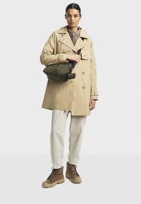Trench beige sopra un pile grigio, abbinato a pantaloni bianco sporco e stivali marroni. Tiene una borsa trapuntata verde oliva con una tracolla nera.