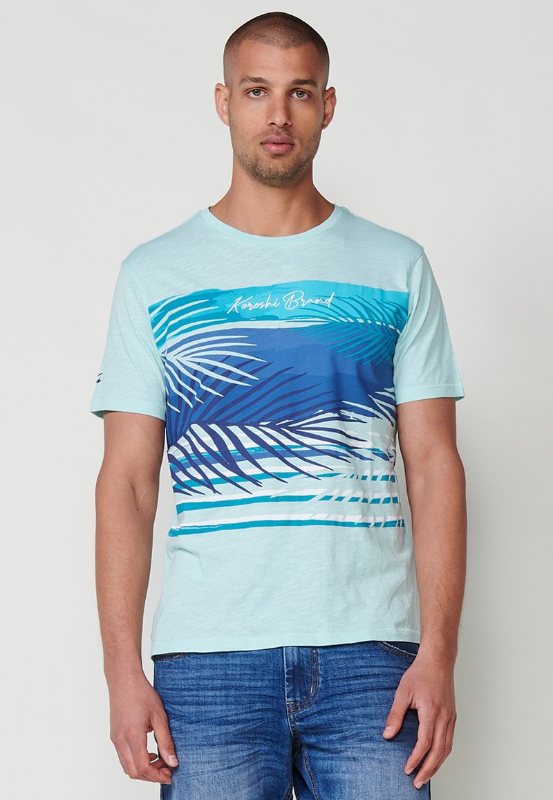 Ljusblå t-shirt med kort ärm som har en grafisk design av blå palmblad och ränder, tillverkad av ett mjukt tyg med en avslappnad passform.
