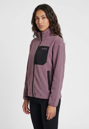 Femme aux longs cheveux ondulés portant une veste polaire mauve avec des poches zippées noires, debout sur un fond blanc uni.