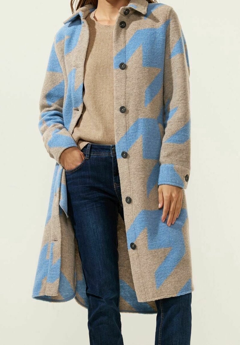 Lång, beige kappa med blå zigzag-mönster. Tillverkad av ett mjukt tyg, har en kragen och svarta knappar. Tillsammans med en ljusbrun tröja och jeans.