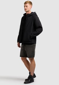 Jeune homme portant une veste à capuche noire, un short en denim gris foncé et des baskets montantes noires, debout les mains dans les poches.