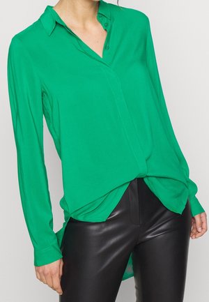 Vrouw draagt een felgroene blouse met lange mouwen en knopen, ingestopt in zwarte leren broek, tegen een effen achtergrond.