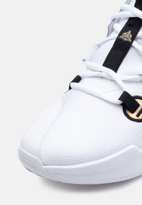 Białe buty sportowe z teksturowaną siateczką na górze, czarnymi sznurowadłami, złotym akcentem z logo oraz zaokrągloną białą gumową podeszwą.