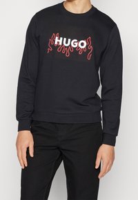 Felpa nera girocollo in cotone con maniche lunghe. Presenta il testo "HUGO" in bianco e motivi di fiamme rosse. Polsini e orlo a costine.