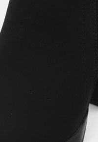 Czarny botek za kostkę wykonany z fakturowanego materiału o gładkim wykończeniu, z okrągłym kształtem, subtelnym przeszyciem oraz niskim obcasem.